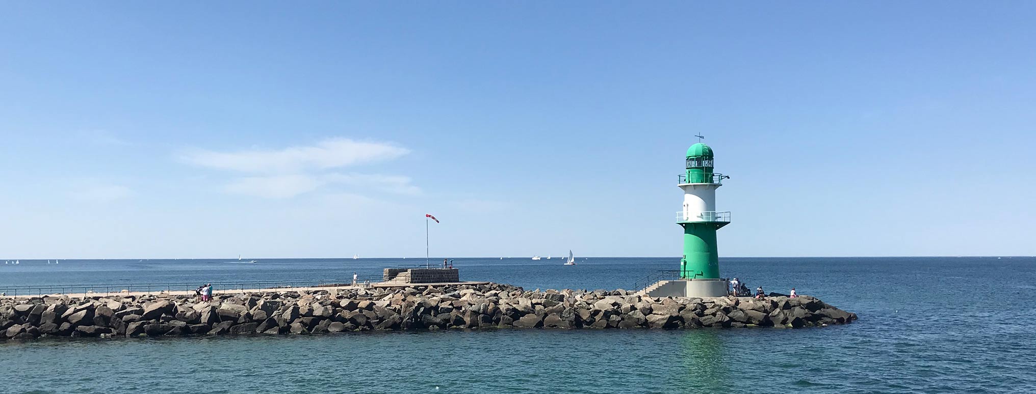 Grün-weißer Leuchtturm an der Mole mit Blick auf das offene Meer und Segelboote im Hintergrund.