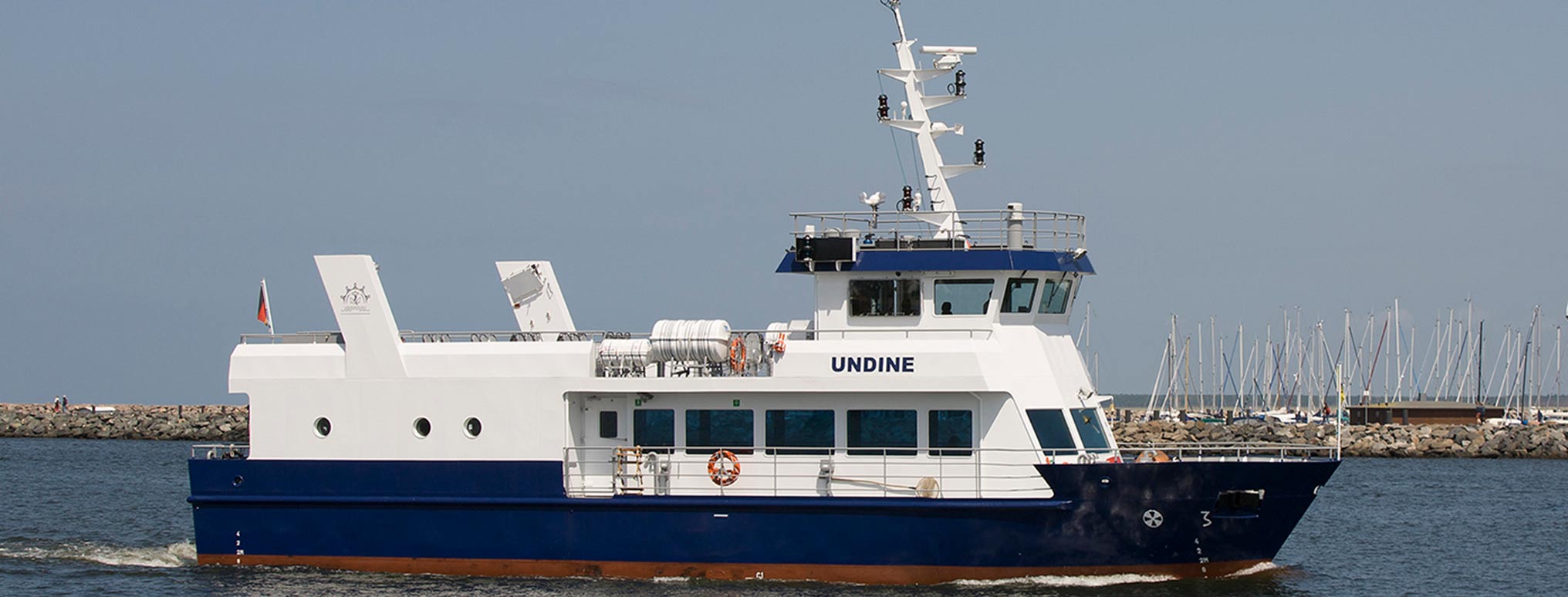 Das Schiff 'Undine' auf dem Wasser, bereit für eine Seebestattung, mit Hafen und Segelbooten im Hintergrund.