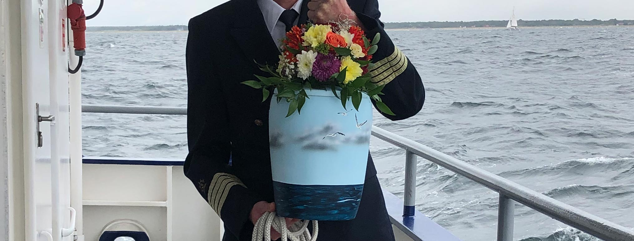 Kapitän in Uniform hält eine maritime Urne mit Blumen während einer Seebestattung auf hoher See.