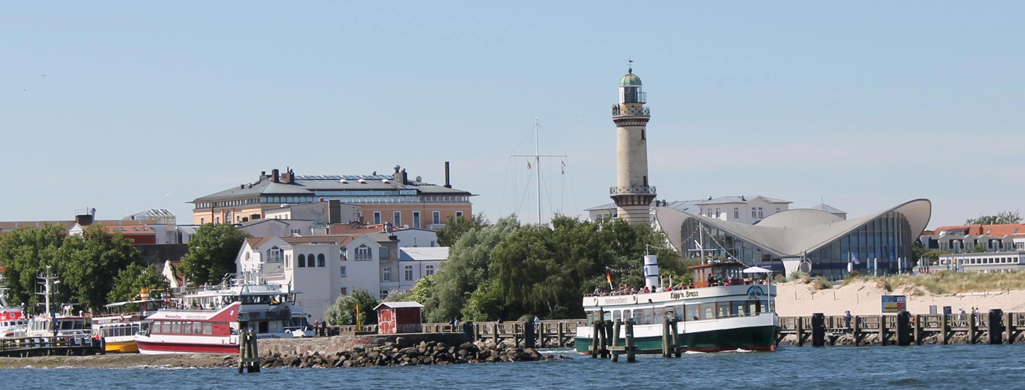 Hafenansicht von Warnemünde mit historischem Leuchtturm und moderner Architektur im Hintergrund.